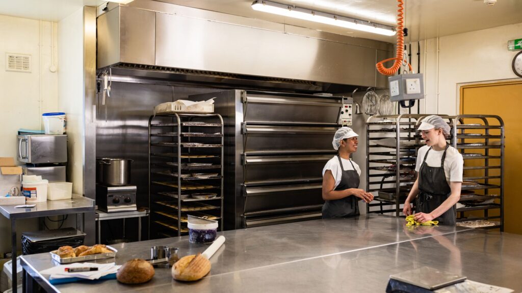 Bakery staff cleaning a large stainless steel workbench in a food production kitchen