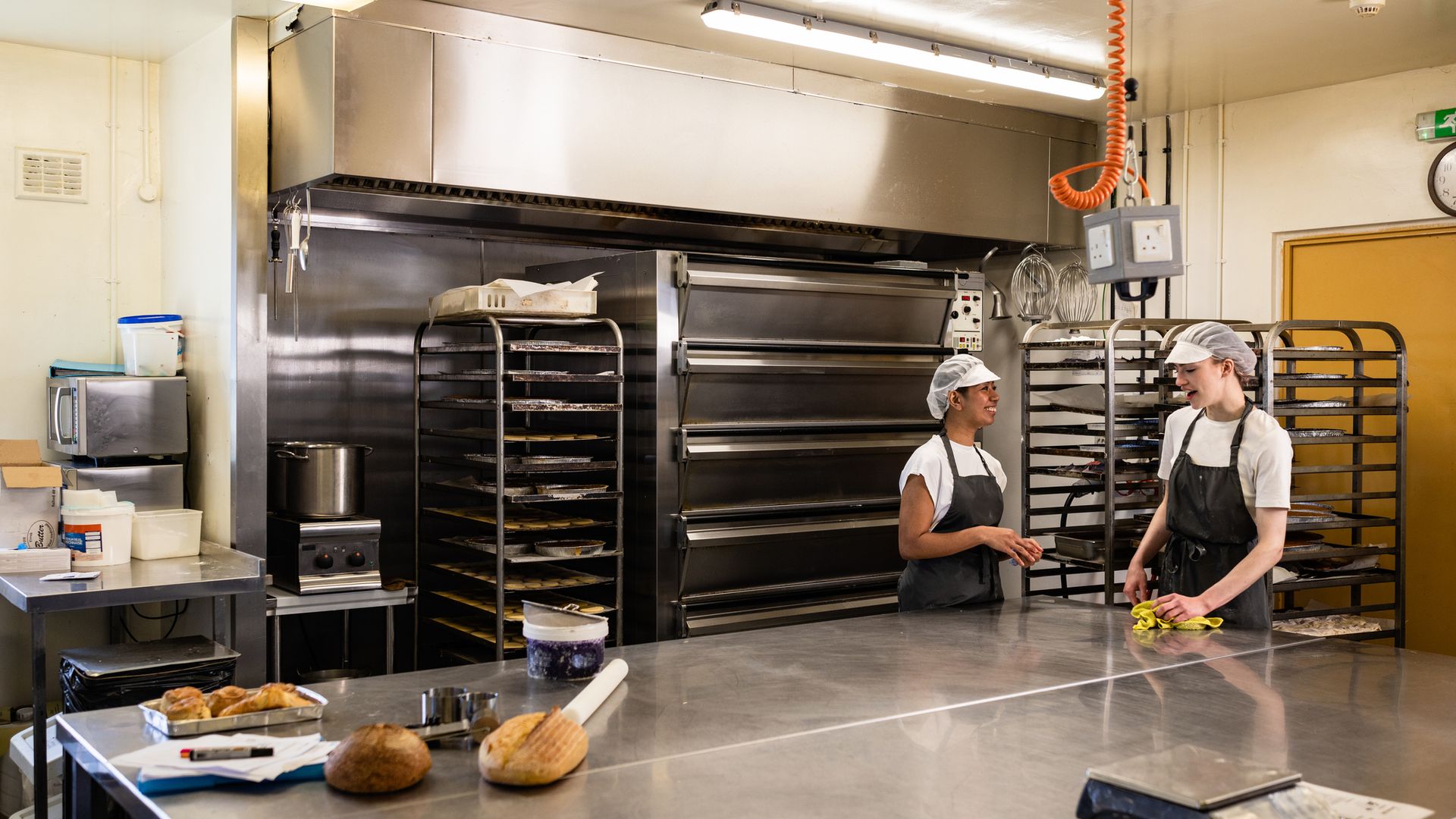 Bakery staff cleaning a large stainless steel workbench in a food production kitchen